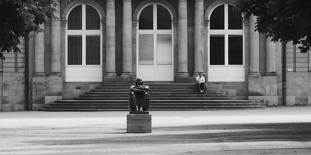 a statue in a plaza, person seated on steps, talking on phone in the background